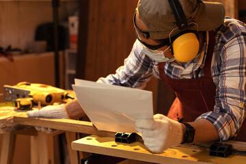 Carpenter wearing safety ear muff with paper drawing while working in carpentry workshop.