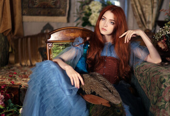 Young red haired woman in long blue dress holding blue peacock in hands in vintage room. Fantasy...