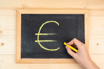 A woman's hand draws the euro symbol in yellow chalk on a black blackboard in a frame
