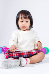 Studio shot of little cute short black hair Asian baby girl daughter model in casual plaid skirt sitting on floor smiling laughing playing with colorful round balls toy alone on white background