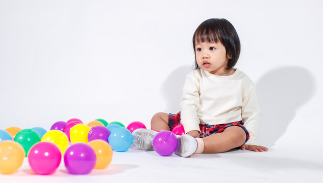 Studio shot of little cute short black hair Asian baby girl daughter model in casual plaid skirt sitting on floor smiling laughing playing with colorful round balls toy alone on white background