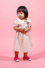Portrait studio shot little cute Asian kindergarten preschooler girl daughter model in casual long dress and shoes standing look at camera holding hundred dollars banknote in hands on pink background