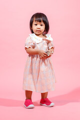Portrait studio shot little cute Asian kindergarten preschooler girl daughter model in casual long dress and shoes standing look at camera holding hundred dollars banknote in hands on pink background