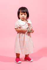 Portrait studio shot little cute Asian kindergarten preschooler girl daughter model in casual long dress and shoes standing look at camera holding hundred dollars banknote in hands on pink background