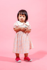 Portrait studio shot little cute Asian kindergarten preschooler girl daughter model in casual long dress and shoes standing look at camera holding hundred dollars banknote in hands on pink background