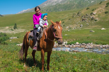 Mother with child are sitting on horse back with green mountains background.