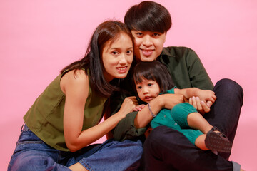 Portrait closeup studio shot of young happy Asian family father mother and little cute baby girl daughter sitting on floor together smiling look at camera on pink background