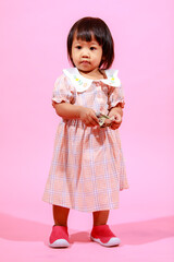 Portrait studio shot little cute Asian kindergarten preschooler girl daughter model in casual long dress and shoes standing look at camera holding hundred dollars banknote in hands on pink background