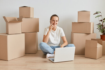 Positive woman wearing white T-shirt sitting on the floor near cardboard boxes with personal stuff,...