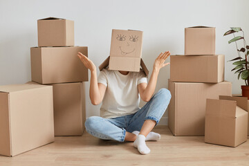 Indoor shot of unknown anonymous woman wearing white T-shirt sitting on the floor near cardboard boxes with personal pile, posing with carton box on her head, spreading hands aside.