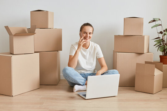 Positive woman wearing white T-shirt sitting on the floor near cardboard boxes with personal stuff, talking phone, looking at camera, expressing positive emotions.