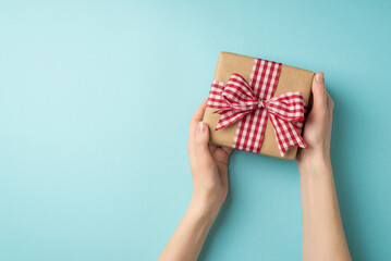 First person top view photo of valentine's day decorations young person's hands giving kraft paper giftbox with checkered ribbon bow on isolated pastel blue background with blank space