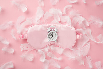 Top view photo of white alarm clock on pink silk sleeping mask and feathers on isolated pastel pink background