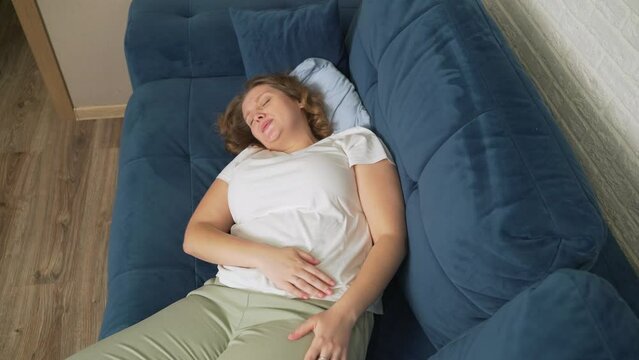 Woman in white T-shirt is lying on blue sofa and holding stomach with hand. She strokes stomach with hand so that it does not hurt. Menstruation, pregnancy, diarrhea, gluttony. There is pain on face
