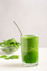 Homemade green organic spinach smoothie or juice served in drinking glass with straw and leaves ingredient in bowl on white wooden table. Healthy dieting concept. Vertical image with copy space