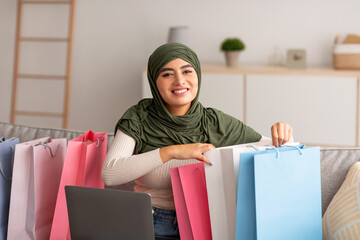 Happy Arab woman in hijab sitting on couch with laptop and bright shopper bags, buying goods online at home