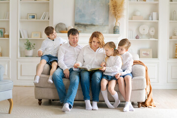 Happy family reading book at home. Mom and dad and young children sit together on a cozy sofa in a new large, bright living room with white walls, furniture and shelves in the background.