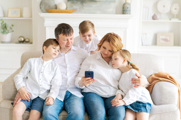 Caucasian family sitting on sofa in living room at home. Mother father little daughter and two sons using a smartphone are watching an entertainment program, videos online, talking on a video call