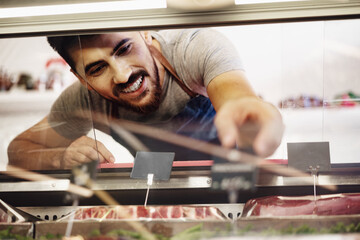 Young man butcher arranging meat products in display case of butcher shop