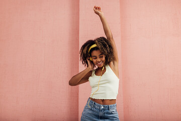 Smiling woman raising hand up while listening to music and dancing at a pink wall