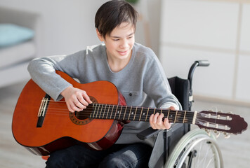 Cheerful impaired teen boy in wheelchair learning how to play guitar and singing indoors