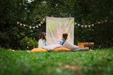 Back view of sisters spending time together and watching cimema at backyard. Open air cinema. Backyard Family outdoor movie night with kids. DIY Screen with film. Summer outdoor  activities