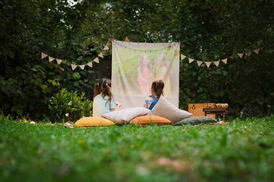 Back View Of Sisters Spending Time Together And Watching Cimema At Backyard. Open Air Cinema. Backyard Family Outdoor Movie Night With Kids. DIY Screen With Film. Summer Outdoor  Activities