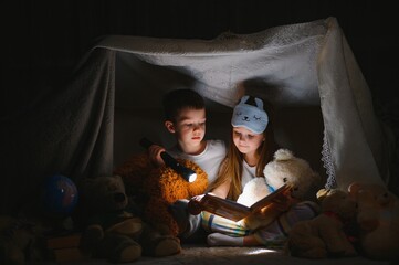 Obraz na płótnie Canvas Siblings sit in a hut of chairs and blankets. Brother and sister reading book with flashlight at home