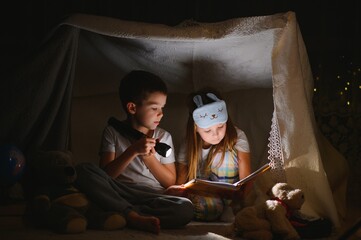 Fototapeta na wymiar Two children with flashlight read a book under a blanket as a tent