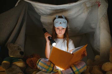 Obraz na płótnie Canvas Little girl are reading a book with flashlights in tent. Happy girl playing at home. Funny lovely kid having fun in room.