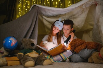 Obraz na płótnie Canvas Two children with flashlight read a book under a blanket as a tent