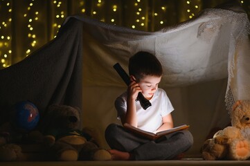 boy reading book with flashlight in tent at night - obrazy, fototapety, plakaty