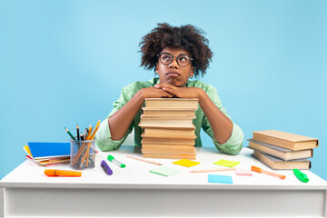 Sad african american male student sitting at table and leaning on stack of books, doesn't want to study, blue background