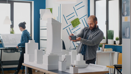 Man engineer using smartphone to work on building model and layout in architectural office. Architect working with mobile phone to design construction print plans and urban project.