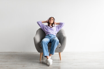 Obraz premium Full length of peaceful young lady in headphones relaxing in armchair, listening to music against white studio wall