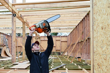 young guy holds chainsaw over his head at arm's length and looks at it. Portrait of worker on background of wooden frame house under construction