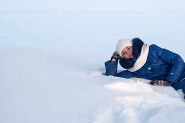 Fototapeta na wymiar beautiful girl in warm blue coat and white hat and scarf is lying on snow. Teenage girl walks on frosty morning and enjoys nature. There is lot of snow around