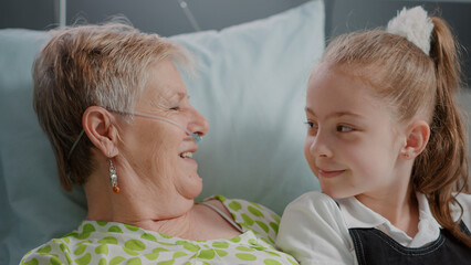 Close up of senior woman with illness enjoying visit from litte girl in hospital ward bed. Aged patient with sickness having fun with niece visiting to give support and assistance.