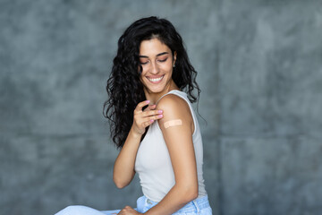 Fototapeta premium Young Caucasian woman showing adhesive bandage on her arm, getting vaccinated against covid-19 on grey background