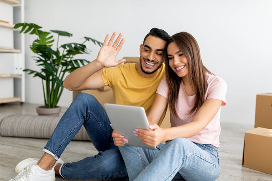 Young multinational couple chatting with friends or family online on tablet pc in new apartment. Relocation concept