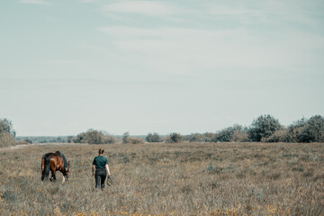 Caucasian young woman and free bay horse walking through pasture, back view.