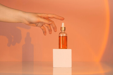 Dropper glass Bottle Mock-Up next to geometric shapes and female hand in the light of the setting sun. Oily drop falls from cosmetic pipette on pink and beige background. Cosmetology, beauty concept.