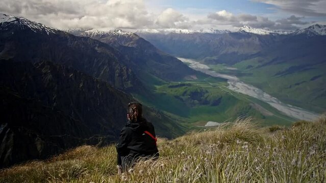 Girl looking over a beautiful green valley with mountains with a river running through on a bright sunny day on a grassy hill, drifting handheld wide shot. New Zealand.