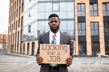 Young black businessman holding cardboard sign with the text LOOKING FOR A JOB, lost his job due to...