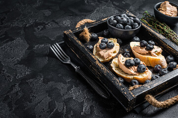 Foie gras toasts, duck liver pate and fresh blueberry in wooden tray. Black background. Top view. Copy space - obrazy, fototapety, plakaty