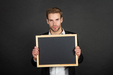 Serious teacher in formal suit holding empty chalkboard copy space dark background, school