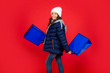 cheerful kid in puffer jacket and hat. teen girl after shopping on red background.