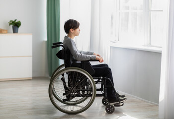 Fototapeta na wymiar Unhappy impaired teenage boy sitting in wheelchair, looking out window, feeling desperate and alone at home
