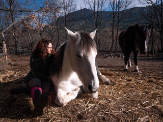 Side view of female in green polar fleece jacket sitting by the side of a white pony with other...