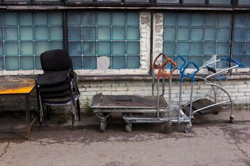 piled old dirty dusty office chairs, stacked cargo trolleys and a rusty weathered yellow metallic table standing outside on a soviet blue glass bricks windows background